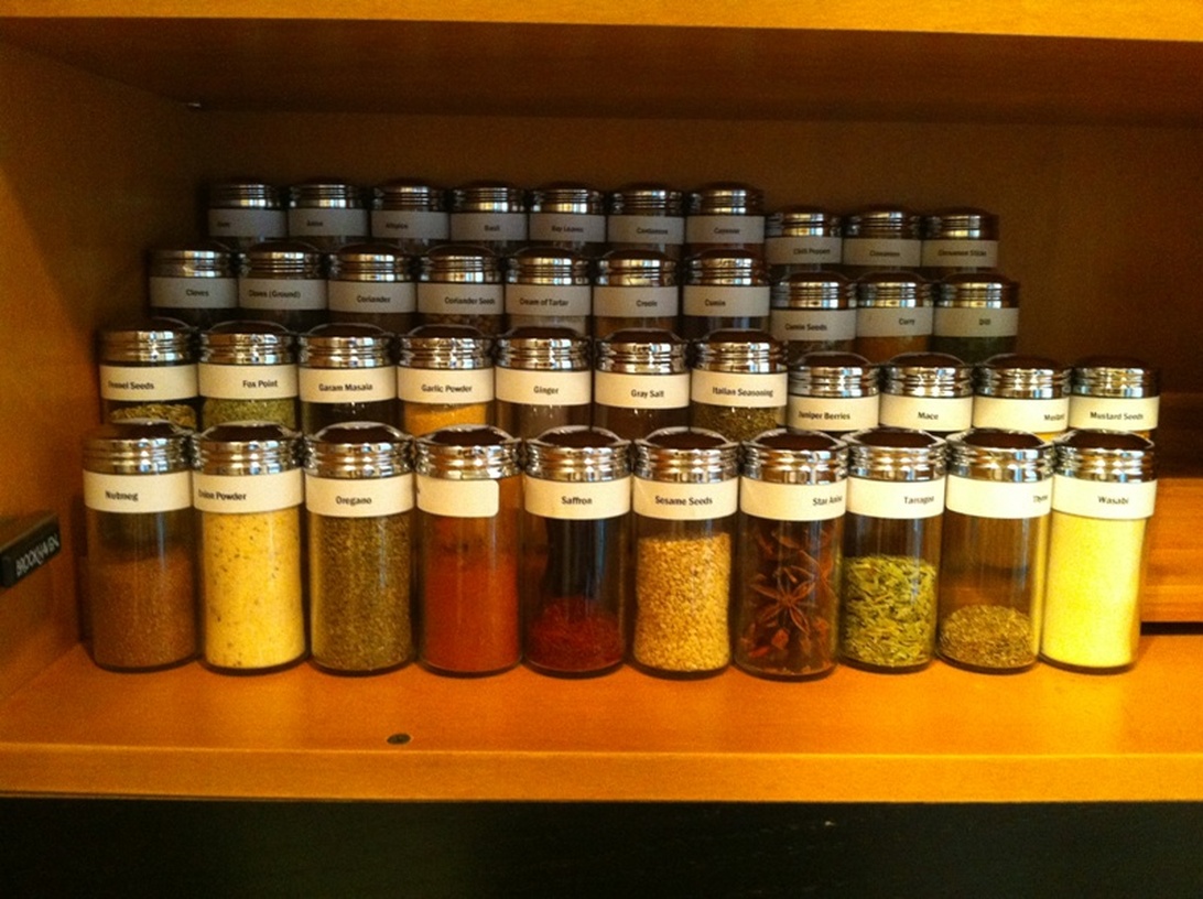 En välorganiserad samling av matchande glasburkar med kryddor som fänkålsfrön, five spice, garam masala, vitlökspulver, ingefära, stjärnanis och saffran, tydligt märkta och uppradade på kökshyllor i trä.